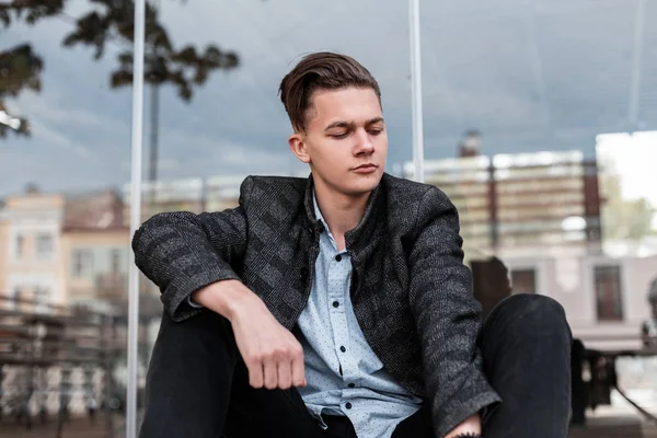 Hombre joven europeo guapo en chaqueta a cuadros vintage gris en camisa de moda en jeans con peinado elegante sentado cerca de un moderno café de verano de vidrio en la ciudad. Guymodel lindo es relajante al aire libre . — Foto de Stock