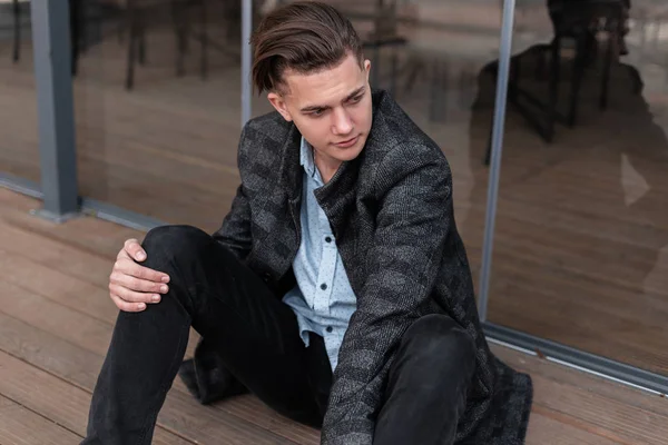 Joven hombre guapo con un peinado de moda en una elegante chaqueta a cuadros gris en una camisa en jeans vintage se sienta al aire libre cerca de puertas de vidrio. Un tipo atractivo descansando cerca del café de verano. Moda . — Foto de Stock