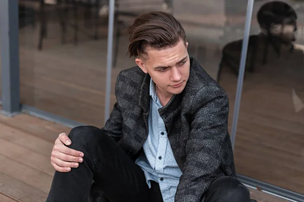 Μοντέρνα δροσερό νεαρό άνδρα με ένα μοντέρνο χτένισμα σε κομψά ρούχα χαλαρώνει καθισμένος κοντά στις σύγχρονες γυάλινες πόρτες κοντά στο καλοκαιρινό καφέ σε εξωτερικούς χώρους. Ευρωπαϊκό μοντέλο τύπος αναπαύεται στην πόλη. — Φωτογραφία Αρχείου