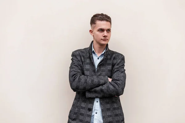 Joven hombre fresco con un peinado elegante en una chaqueta a cuadros gris largo en una camisa de moda está de pie cerca de una pared blanca en la calle en un día de primavera. Lindo chico agradable está disfrutando de un paseo . — Foto de Stock