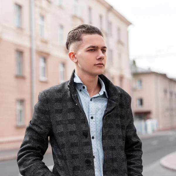 Молодий вродливий європейець у старовинному сірому штампованому куртці в стильній сорочці зі стильною зачіскою в місті біля будинків. Привабливий сучасний хлопець їде по вулиці.. — стокове фото