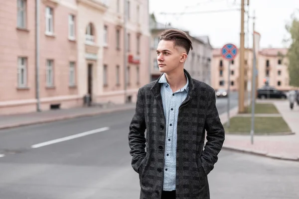 Bonito joven guapo con un peinado de moda en una chaqueta a cuadros de lujo gris en una camisa camina por la ciudad cerca de edificios antiguos en un día de primavera. Cool chico moderno . — Foto de Stock