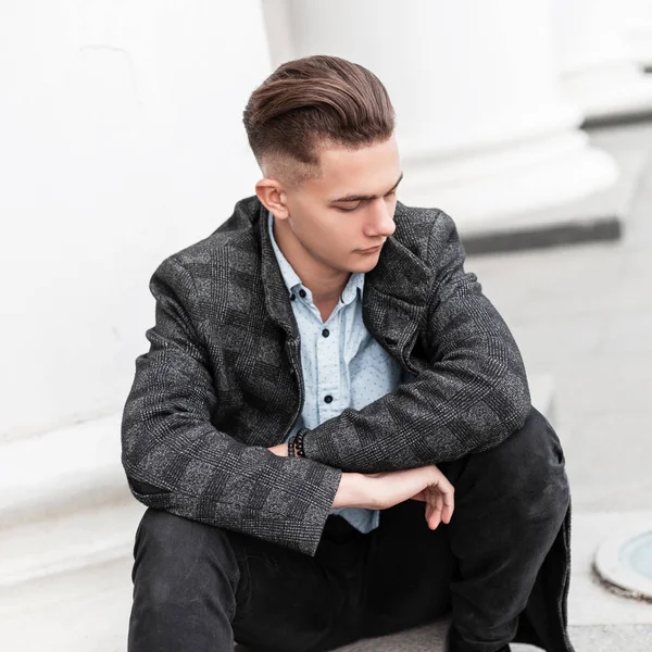 Joven hombre moderno en elegante chaqueta gris a cuadros en una camisa elegante en vaqueros de moda con un peinado de moda está descansando sentado en los escalones cerca de un edificio vintage en la ciudad. Buen tipo modelo . — Foto de Stock