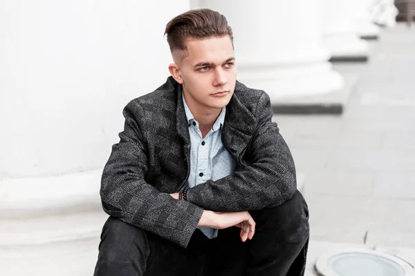 Modern ung man i elegant rutig grå jacka i en snygg skjorta i trendiga jeans med en fashionabel frisyr vilar sitter på trappan nära en Vintage byggnad i staden. Urban Guy-modell. — Stockfoto
