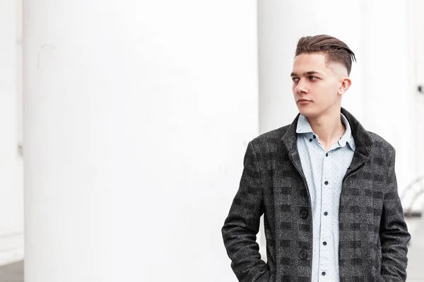 Hombre joven guapo urbano con una camisa elegante en una chaqueta a cuadros vintage gris con un peinado de moda se encuentra cerca de las columnas blancas de un edificio moderno de la ciudad. Negocio buen tipo al aire libre . — Foto de Stock