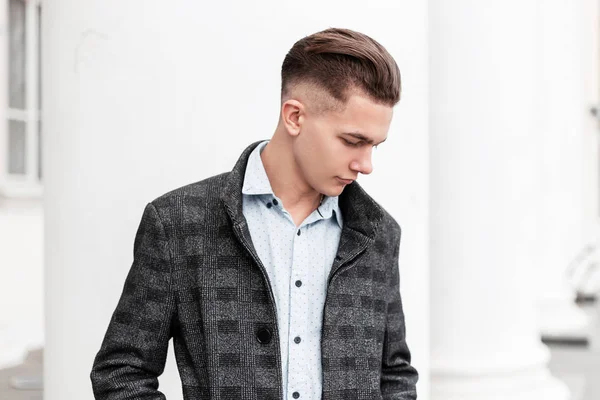 Business young man in a stylish plaid gray jacket with a fashionable hairstyle in a classic shirt is standing near a vintage building on the street in the city. Nice guy is a model for a walk. — Stock Photo, Image