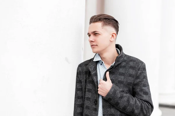 Moderne gars à succès avec une coiffure élégante dans des vêtements à la mode posant debout près d'un bâtiment vintage blanc dans la ville. Homme d'affaires attrayant sur une promenade dans le jour du printemps. Vêtements homme tendance . — Photo