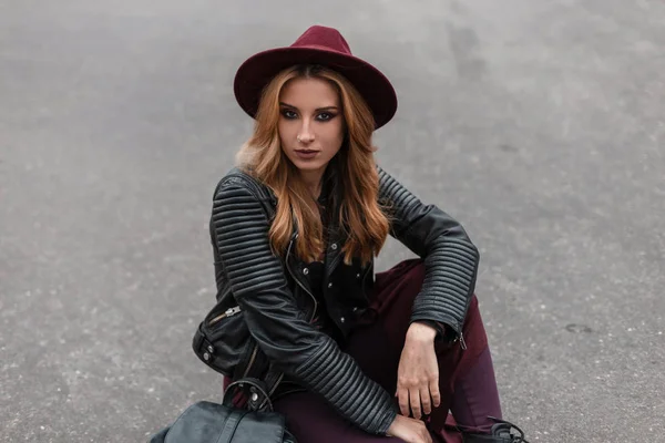 Beautiful pretty young woman in a purple hat in a fashionable leather jacket in vintage pants with a trendy backpack sits on the asphalt outdoors on a spring day. Modern cute red-haired girl resting. — Stock Photo, Image