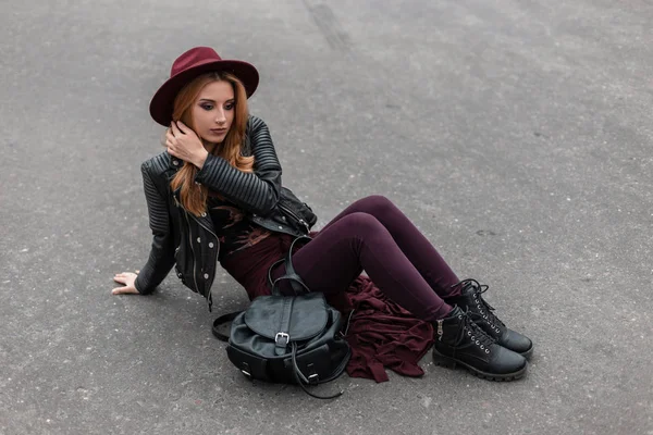 Mujer joven de pelo rojo bonito con un sombrero vintage en una chaqueta negra en pantalones de moda con una mochila de cuero de moda disfruta del resto sentado en el asfalto. Urbano hermosa chica relaja al aire libre . —  Fotos de Stock