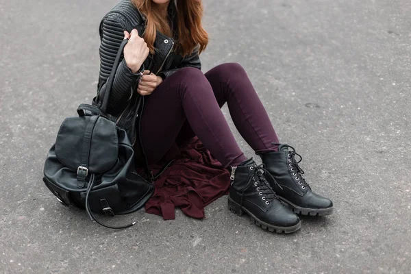Mujer joven en una chaqueta de estilo negro en pantalones de color púrpura vintage en botas de cuero de moda con una mochila de cuero se sienta en el asfalto. Nueva colección estacional de zapatos de mujer de moda. Primer plano . —  Fotos de Stock