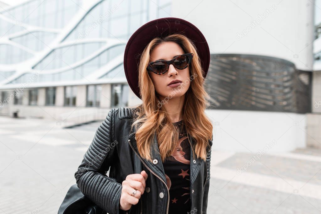 Portrait of a redhaired attractive young hipster woman in an elegant hat in trendy sunglasses in a black leather jacket with a backpack with an earring in nose outdoors in the city. Modern urban girl.