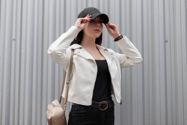 Bir metal duvara yakın bir şehirde poz şık bir siyah beyzbol şapkası siyah kot pantolon bir t-shirt moda altın sırt çantası ile güzel genç esmer kadın. Avrupalı kız