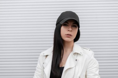 Modern bir metal duvara yakın şık bir beyaz deri ceket şık bir siyah beyzbol şapkası oldukça çekici esmer genç kadın Portresi. Güzel çekici kız modeli açık havada. Moda.