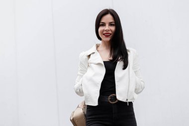 Kot pantolon bir T-shirt şık bir deri ceket şirin bir gülümseme ile güzel mutlu genç kadın bir bahar parlak bir günde beyaz bir binanın yakınında duruyor. Pozitif kız modeli tatil sahiptir.