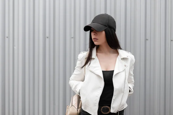 Jolie jeune femme brune américaine vêtue d'une veste en cuir blanc tendance avec une élégante casquette de baseball noire dans un T-shirt avec un sac à dos en or posant près d'un bâtiment en métal. Une fille sexy moderne. Style de rue . — Photo