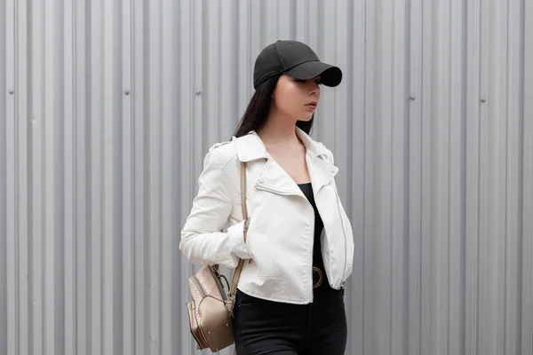 Mulher muito atraente em um boné de beisebol preto na moda em uma jaqueta de couro branco elegante em jeans com uma mochila de ouro vintage fica no dia de primavera perto de um edifício de metal cinza. Menina bonita . — Fotografia de Stock