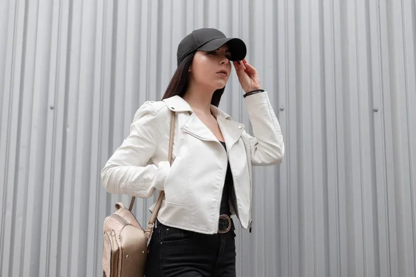 Moderní atraktivní mladá žena ve stylové baseballové čepici ve starožitném bílém koženém kabátku v černých džínách s módním batohu u moderní kovové budovy ve městě. Americká dívka venku. — Stock fotografie