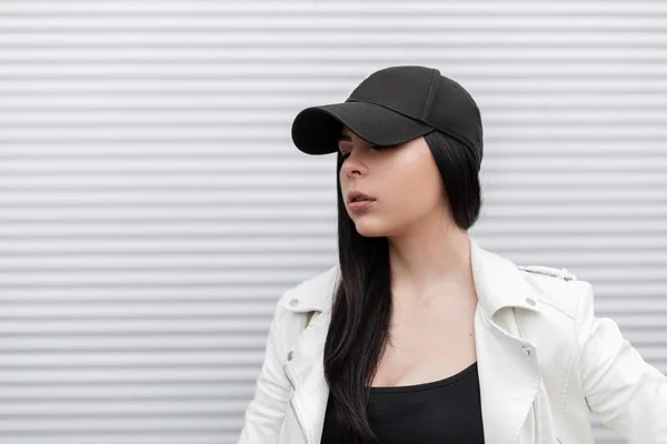 세련된 검은 색 야구 모자를 쓰고 세련된 흰색 가죽 재킷을 입고 야외에서 실버 메탈 월 근처의 티셔츠를 입은 예쁜 귀여운 젊은 여성. 미녀. 여성의 얼굴의 야외 초상화. — 스톡 사진