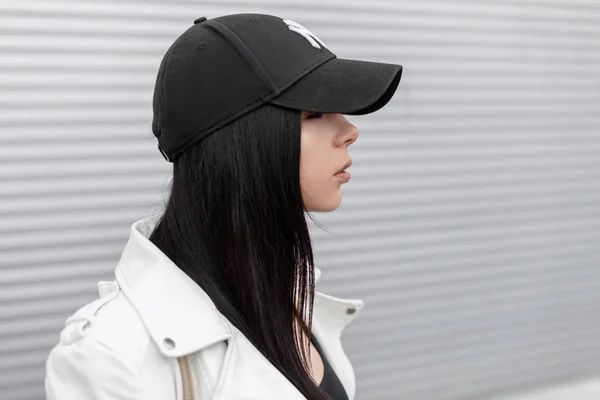 Porträtt av ganska ung vacker kvinna i en snygg vit skinnjacka i en fashionabel Svart keps nära en metall vägg i staden. Attraktiv brunett tjej. Side View. — Stockfoto