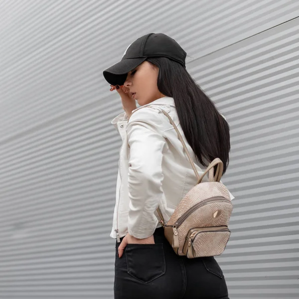 Modaya uygun bir beyzbol şapkası bir vintage sırt çantası ile şık bir beyaz deri ceket Kentsel güzel genç kadın bahar gününde bir metal binanın yakınında şehir etrafında yürüyor. Şık kız. Geri görünüm. — Stok fotoğraf