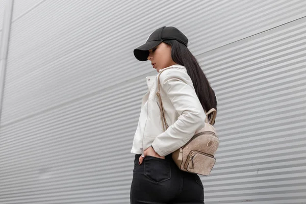 Americká mladá žena ve stylové čepici s módním zlatým batohu v bílém koženém kabátku, které cestovala po ulici u kovové zdi. Atraktivní dívčí módní model. Styl ulice mládeže. — Stock fotografie