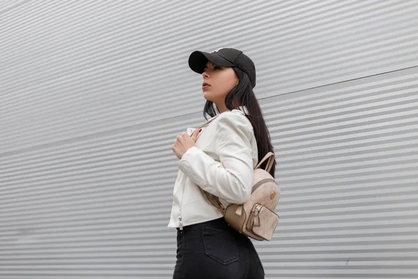 Mulher muito bonita em uma jaqueta de couro branco na moda em jeans pretos elegantes com uma mochila de ouro na moda anda pela cidade perto de um edifício de metal moderno. Menina urbana atraente viagens . — Fotografia de Stock