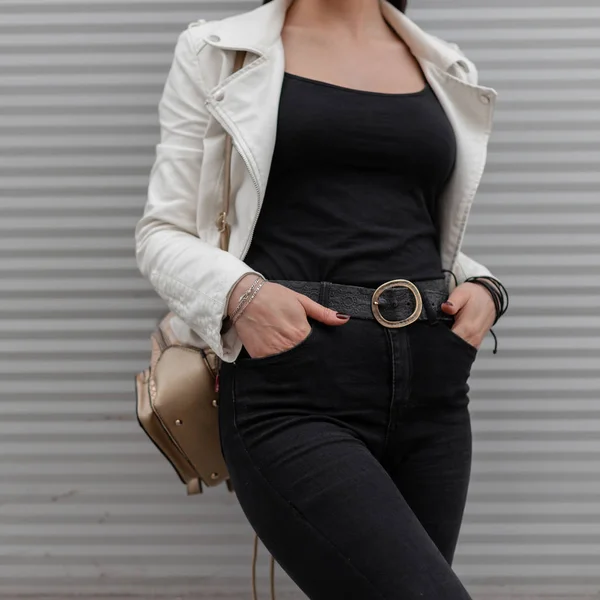 Young woman in white leather jacket in a t-shirt in black jeans with a stylish golden backpack poses near the modern wall. Details of everyday look. Fashionable women's modern clothing. Close-up. — Stock Photo, Image