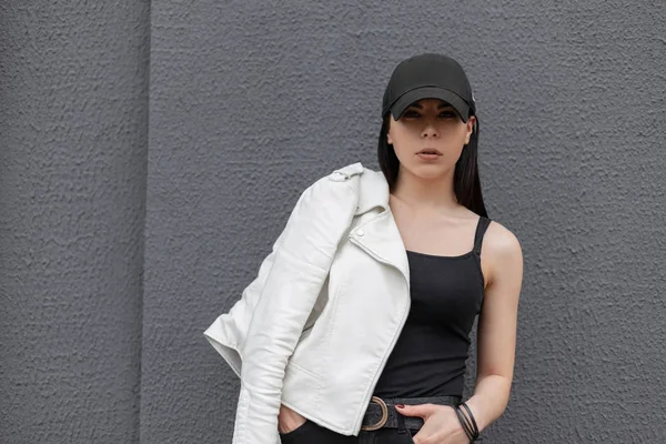 Νεαρή όμορφη γυναίκα με μαύρο T-shirt σε λευκό κομψό δερμάτινο μπουφάν σε μοντέρνο μαύρο καπέλο ποζάρει κοντά σε ένα γκρι κτίριο στην πόλη μια ανοιξιάτικη μέρα. Μοντέρνο ελκυστικό γυναικείο μοντέλο. Αμερικάνικο στυλ. — Φωτογραφία Αρχείου
