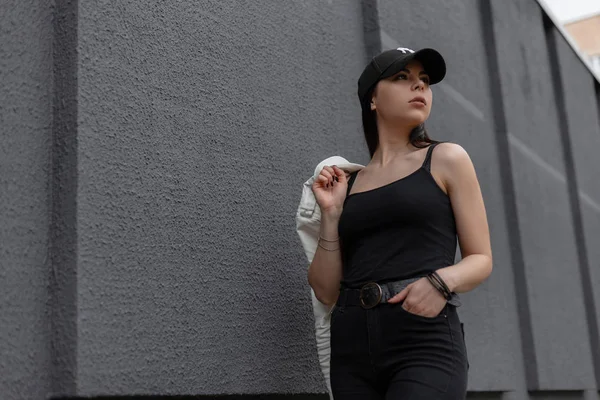Mooie jonge brunette vrouw in een zwart Vintage t-shirt in een modieuze zwarte pet in stijlvolle zwarte jeans met een witte leren jas in de buurt van een grijs gebouw in de stad. Schattig meisje model op een wandeling. — Stockfoto