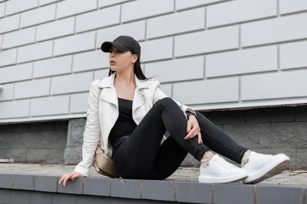 Jovem americana em uma jaqueta de tênis de couro em jeans pretos em um boné de beisebol elegante com uma mochila senta-se em uma telha perto de um edifício vintage na cidade. Menina bonita relaxa ao ar livre . — Fotografia de Stock