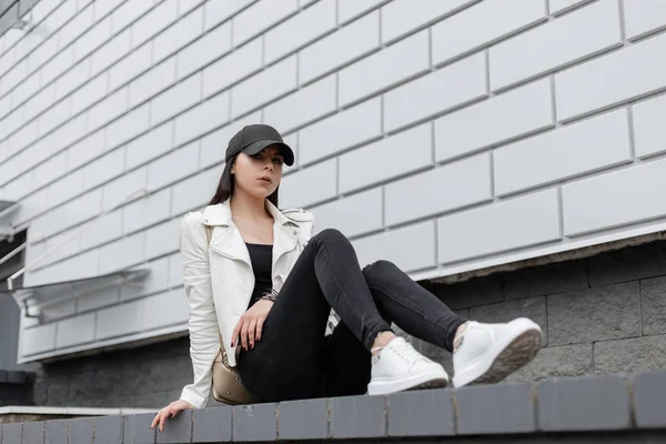 Mooie moderne jonge vrouw in zwarte stijlvolle Cap in een leren jas in jeans in modieuze sneakers met een gouden rugzak rusten buitenshuis zittend op de tegel in de buurt van het gebouw. Mooi meisje ontspant. — Stockfoto