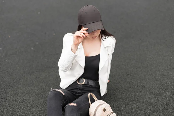 Uma jovem muito moderna e elegante mulher de boné preto em uma jaqueta branca de couro em jeans em uma camiseta com uma mochila dourada está descansando ao ar livre sentada no asfalto. Menina americana relaxa. Estilo de juventude . — Fotografia de Stock