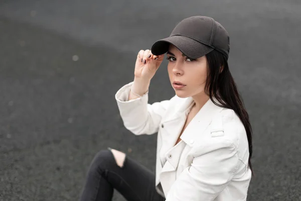 Mooie jonge brunette vrouw met bruine ogen in een stijlvolle Amerikaanse Baseball Cap in een stijlvolle lederen witte jas in gescheurde jeans geniet van de rest zittend op het asfalt op een zomerdag. Schattig meisje. — Stockfoto