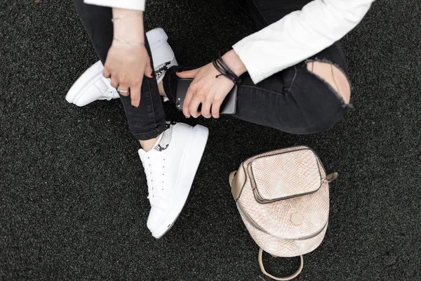 Close-up van een jonge vrouw benen in geripte modieuze zwarte jeans in lederen stijlvolle met sneakers met slang patroon met een trendy gouden rugzak. Stijlvol meisje zit op de stoep in de stad. — Stockfoto