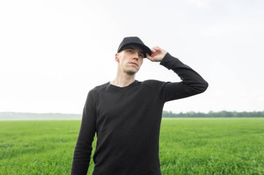 Siyah tişörtlü şık siyah şapkalı genç hoş bir adam yeşil çimenli bir arazide duruyor. Amerikalı erkek modeller şehir dışında tatil yapmayı severler..