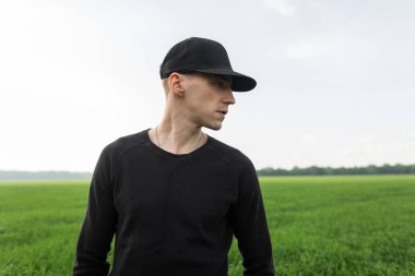 Siyah bir t-shirt bir vintage Amerikan kap yakışıklı genç adam arka planda açık havada mavi bir gökyüzü ve yeşil çim bir alan duruyor. Yakışıklı adam doğada rahatlar.