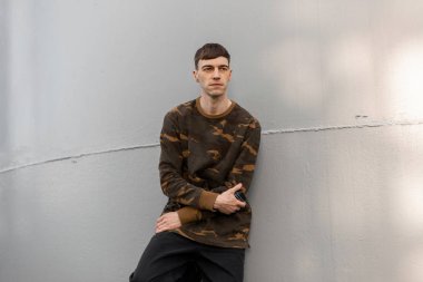 Şık saç stili, modaya uygun bir kamuflaj gömleği ve modaya uygun kot pantolon giyen çekici genç adam, açık havada gri metal bir duvarın yanında güneş ışığının tadını çıkarıyor. Yürüyüş için yakışıklı bir adam. Şık erkek kıyafetleri..