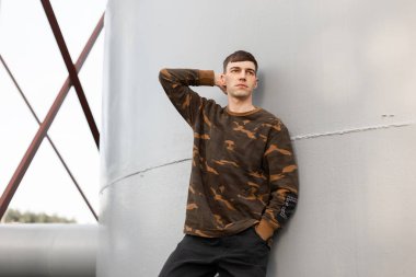 Bir askeri yeşil moda gömlek şık kot moda saç modeli ile Modern genç adam duruyor ve bir yaz gününde gri vintage metal duvar yakınında duruyor. Güzel model adam açık havada rahatlatır.