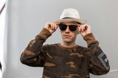 Eski moda gömlekli, klasik şapkalı genç yakışıklı hippi dışarıda metalik gri duvarın yanında duruyor ve şık güneş gözlüklerini düzeltiyor. Amerikan modern erkek modeli.