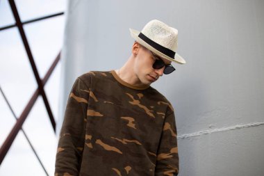 Koyu güneş gözlüklü, şık bir şapka takmış, şık askeri gömlekli genç ve çekici bir hippi gri metal bir binanın yanında duruyor. Yakışıklı, modern bir model. Yaz stili.