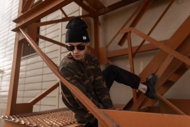 Koyu güneş gözlüğü şık deri spor ayakkabı vintage kot şık bir kamuflaj gömlek Cool genç hipster adam bir demir turuncu merdiven üzerinde oturuyor. Yakışıklı modern adam açık havada dinleniyor