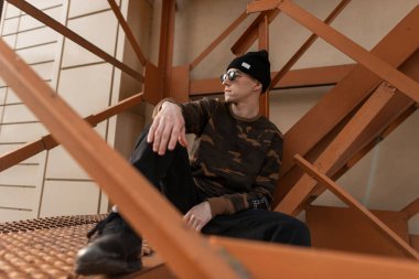 Sokakta demir turuncu merdivenlerde dinlenmiş deri spor ayakkabı moda askeri gömlek şık güneş gözlüğü siyah şapkalı güzel genç hipster adam. Kentsel moda adam açık havada. Erkek giyim