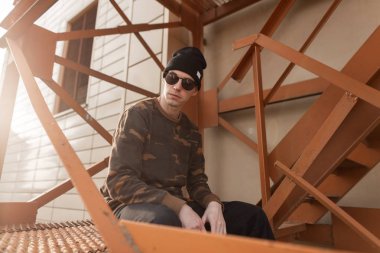 Şık güneş gözlüklü, modaya uygun askeri gömlekli, klasik kot pantolonlu, siyah şapkalı yakışıklı hippi şehirdeki turuncu demir merdivenlerde dinleniyor. Amerikalı çekici adam güneşli bir günün tadını çıkarıyor..