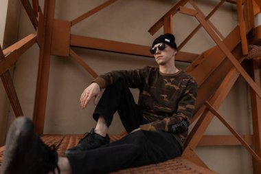 Deri spor ayakkabı koyu güneş gözlüğü vintage siyah şapka şık askeri gömlek Yakışıklı genç hipster adam turuncu demir merdiven oturur. Çekici adam modeli açık havada rahatlatıcı.