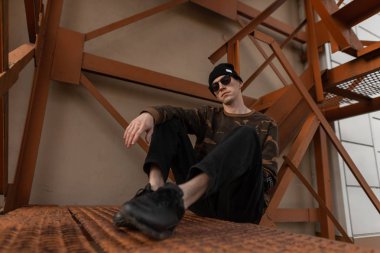 Şık şapkalı, siyah güneş gözlüklü, havalı askeri gömlekli, deri ayakkabılı şık genç hippi şehirdeki metal turuncu merdivenlerde oturuyor. Havalı yakışıklı adam dışarıda..