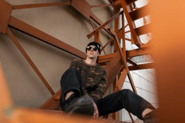 Sokakta demir turuncu merdivenlerde dinlenmiş deri spor ayakkabı yeşil askeri gömlek moda güneş gözlüğü siyah şapkalı modern genç hipster adam. Güzel moda adam açık havada. Erkek giyim