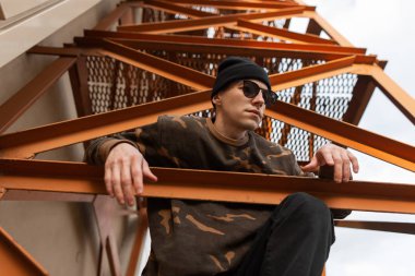 Bir yaz günü açık havada bir demir turuncu merdiven üzerinde oturan güneş gözlüğü kamuflaj gömlek örgü şapka lı güzel genç hipster adam. Modern şık adam modeli. Yakışıklı erkek kıyafetleri.