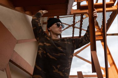 Modaya uygun güneş gözlüğü şık bir askeri gömlek siyah örme şapka lı nice genç hipster adam şehirde bir vintage demir merdiven yakınında pozlar. Parlak güneşli bir günde yürüyen şehirli adam. Sokak stili.