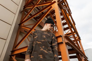 Güneş gözlüğü siyah örme şapka lı modaya uygun bir askeri gömlek güzel genç hipster adam açık havada turuncu bir vintage demir merdiven yakınında duruyor. Şık modern adam bir yaz günü dinleniyor. Sokak stili.