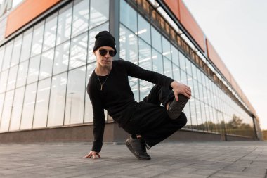 Modaya uygun güneş gözlüğü şık siyah giysiler içinde örme şapkalı modern yakışıklı genç hipster adam cam binanın yakınında şehirde dans ediyor. Şehirli erkek dansçı. Sokak stili. Eğlence.